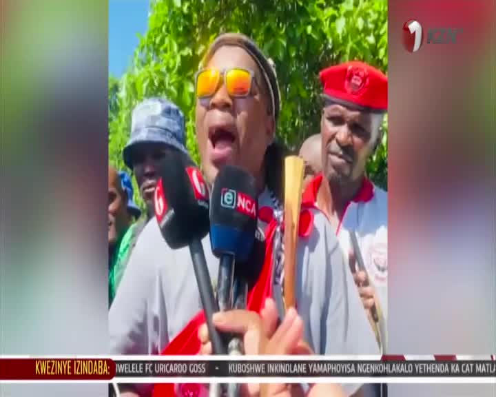 A man in bright orange sunglasses speaks into microphones, his mouth wide open. Beside him, a man in a red beret watches intently, holding a wooden stick. A man in bright orange sunglasses speaks into microphones, his mouth wide open. Beside him, a man in a red beret watches intently, holding a wooden stick.