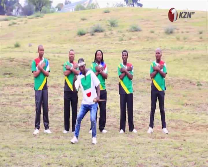 Six men stand in a grassy field, five with arms crossed over their chests, wearing shirts with green, yellow, and red accents. One man in the foreground, in a white shirt with green and red, holds a stick and moves his feet. The KZN TV logo appears in the upper right corner. Six men stand in a grassy field, five with arms crossed over their chests, wearing shirts with green, yellow, and red accents. One man in the foreground, in a white shirt with green and red, holds a stick and moves his feet. The KZN TV logo appears in the upper right corner.