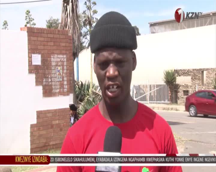 A young man in a red t-shirt and black beanie speaks into a microphone, his front teeth visible. Behind him, a red sedan drives past on a street in South Africa, with a KZN TV logo visible in the upper right corner. A young man in a red t-shirt and black beanie speaks into a microphone, his front teeth visible. Behind him, a red sedan drives past on a street in South Africa, with a KZN TV logo visible in the upper right corner.