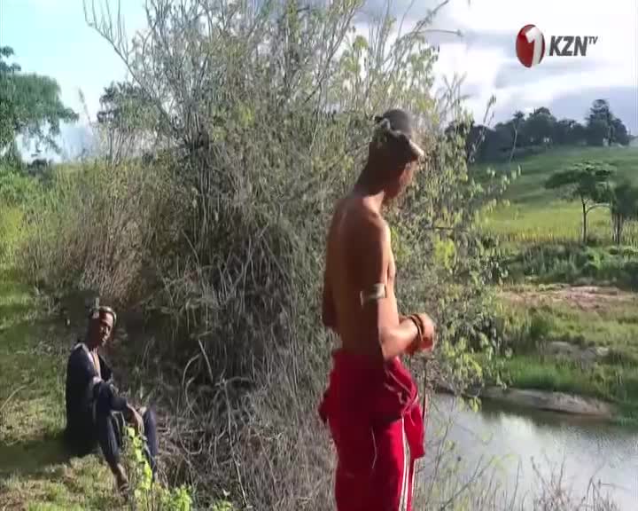 A young man in red track pants stands by the water's edge, his gaze fixed on the scene. Another sits nearby, his dark clothing blending with the shadows of the bush. A young man in red track pants stands by the water's edge, his gaze fixed on the scene. Another sits nearby, his dark clothing blending with the shadows of the bush.