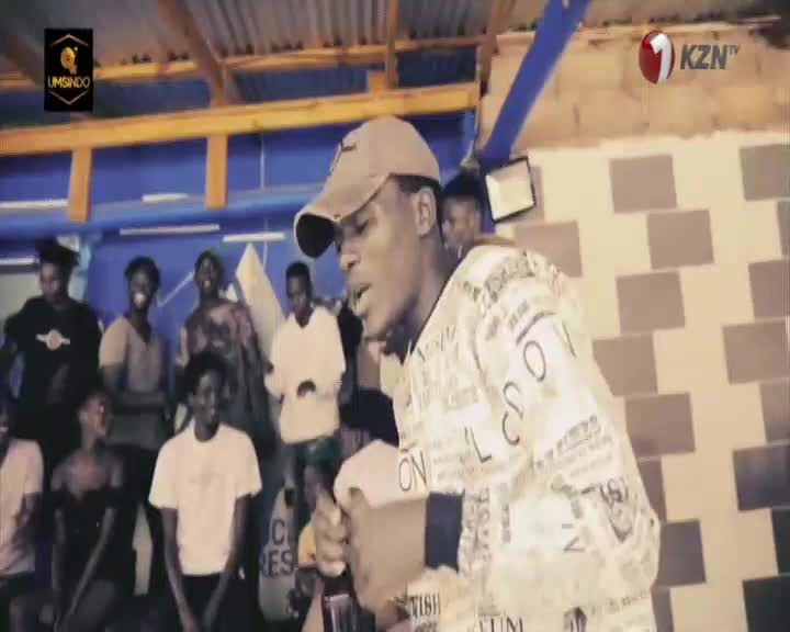 A young man in a patterned shirt and baseball cap raps with intensity, his hands gesturing as a crowd watches in KwaZulu-Natal. The 1KZN TV logo is visible in the corner, indicating the broadcast. A young man in a patterned shirt and baseball cap raps with intensity, his hands gesturing as a crowd watches in KwaZulu-Natal. The 1KZN TV logo is visible in the corner, indicating the broadcast.