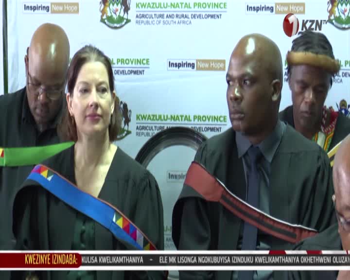A woman in a black robe with a colorful sash looks directly ahead. Beside her, a man in a similar robe and tie glances to his left. Behind them, a banner proclaims "KWAZULU-NATAL PROVINCE AGRICULTURE AND RURAL DEVELOPMENT REPUBLIC OF SOUTH AFRICA." A woman in a black robe with a colorful sash looks directly ahead. Beside her, a man in a similar robe and tie glances to his left. Behind them, a banner proclaims "KWAZULU-NATAL PROVINCE AGRICULTURE AND RURAL DEVELOPMENT REPUBLIC OF SOUTH AFRICA."