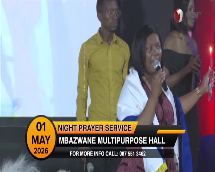 A woman holds a microphone and a lit candle, speaking into it with conviction. Behind her, a man in a mustard-colored shirt stands attentively, and a 1KZN TV logo is visible. A woman holds a microphone and a lit candle, speaking into it with conviction. Behind her, a man in a mustard-colored shirt stands attentively, and a 1KZN TV logo is visible.