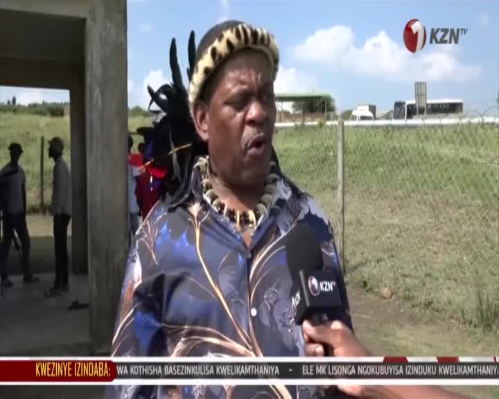 A man in traditional attire speaks into a microphone held by a reporter. Behind him, a few people stand near an open doorway, and beyond a fence, buses travel on a highway. A man in traditional attire speaks into a microphone held by a reporter. Behind him, a few people stand near an open doorway, and beyond a fence, buses travel on a highway.