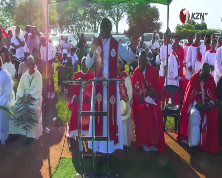A man in a white and red robe stands at a podium, gesturing with his hands. A crowd of people in similar attire fills the background, with vehicles parked behind them. A man in a white and red robe stands at a podium, gesturing with his hands. A crowd of people in similar attire fills the background, with vehicles parked behind them.