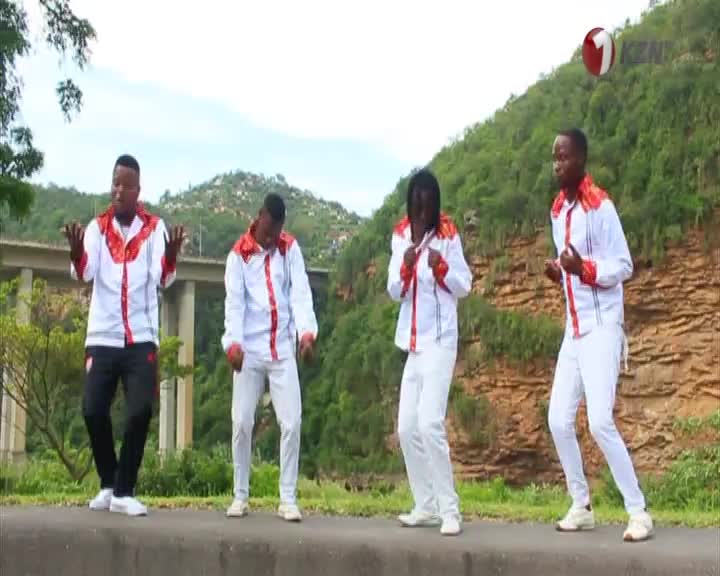 Four young men in matching white and red outfits are dancing by the roadside, with a bridge and a hillside behind them. The 1KZN TV logo is visible in the top right corner.