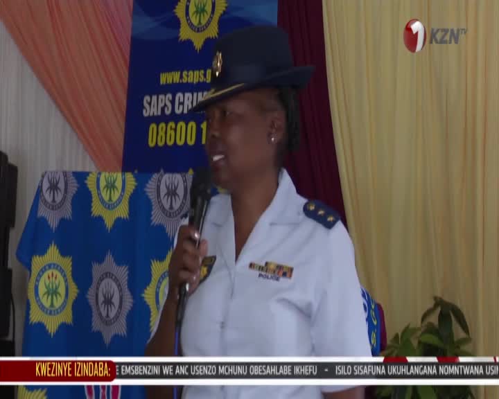 A police officer in uniform speaks into a microphone, her voice carrying over the din of the crowd. Behind her, a banner proclaims "SAPS CRIME LINE" with a phone number. The 1KZN TV logo is visible in the corner, indicating this event is being broadcast live.