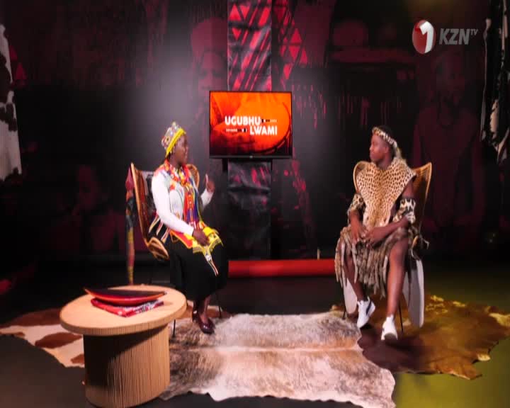 Two people are seated in a studio, one in traditional Zulu attire, facing a screen displaying the 1KZN TV logo. The other person, also in traditional dress, gestures as they speak.