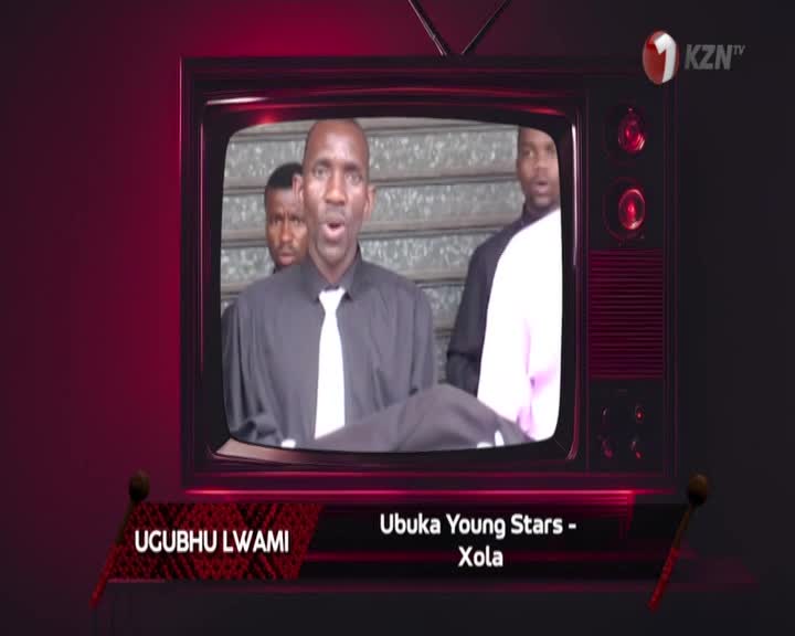 A man in a black shirt and white tie sings with his mouth open, while two other men stand behind him. The KZN TV logo appears in the corner of the screen.