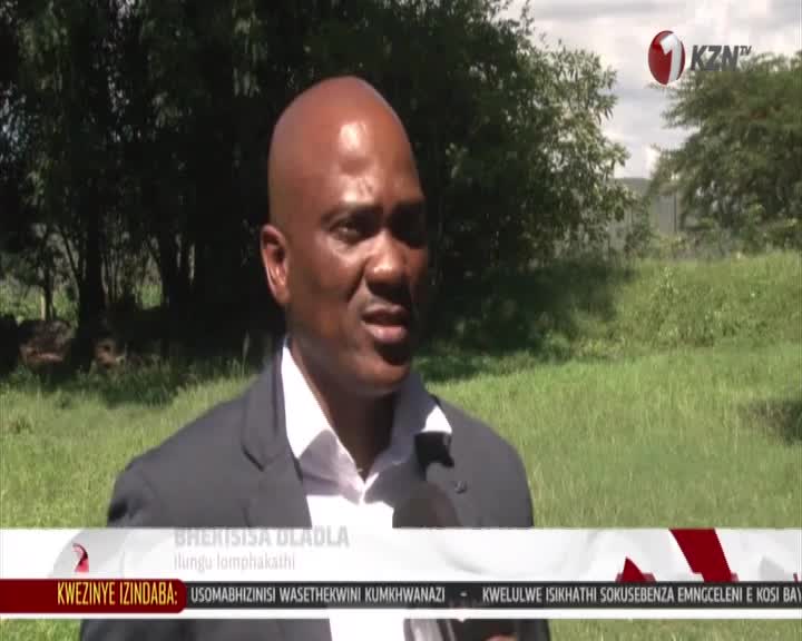 A man in a suit speaks into a microphone, his face illuminated by the sun. Behind him, tall grass sways gently under a cloudy sky.