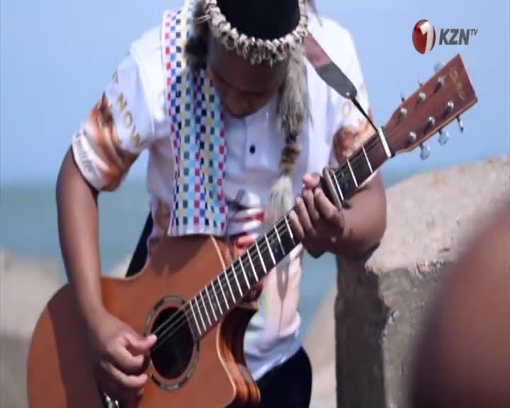 A young man in traditional attire plays an acoustic guitar by the ocean. The KZN TV logo is visible in the corner.