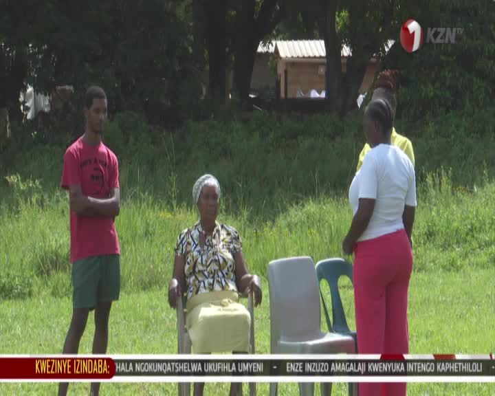 A young man in a red t-shirt stands with his arms crossed, watching an older woman seated in a chair. Another woman in bright pink pants turns away, facing a third person partially visible behind her.