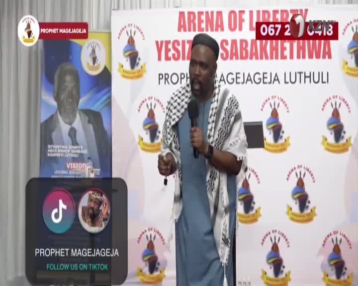 A man in a blue robe and a checkered scarf speaks into a microphone, gesturing with his free hand. Behind him, a banner reads "ARENA OF LIBERTY YESIZWE SABAKHETHWA" in large red letters.