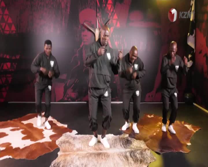 Four men in matching black tracksuits are performing a synchronized dance. They move with their arms bent and hands clasped, their feet tapping on animal hides laid out on the floor. The 1KZN TV logo is visible in the background.