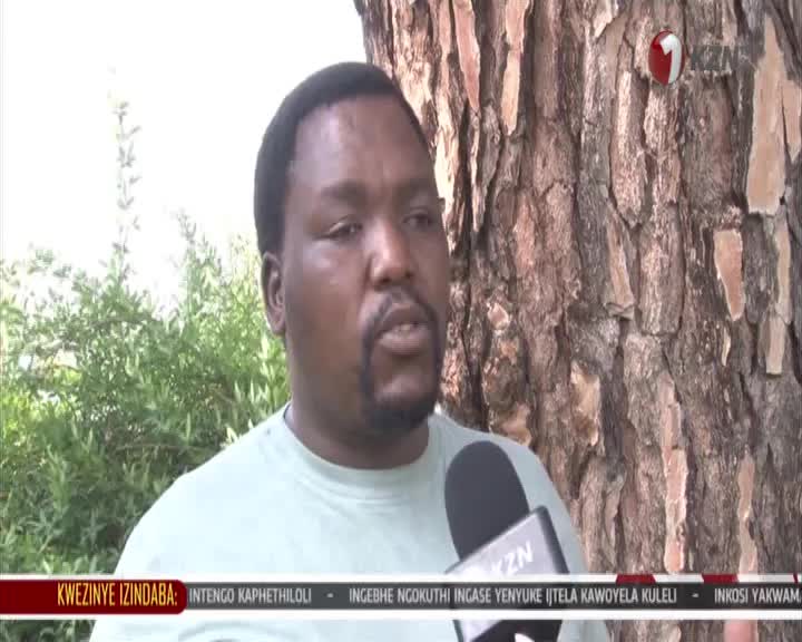 A man speaks into a microphone, his words relayed by 1KZN TV. Behind him, the rough bark of a large tree dominates the frame.