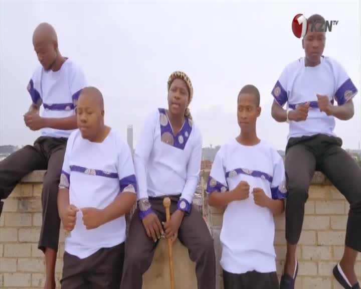 Five men in matching white shirts with blue and purple trim are seated on a brick ledge. One man in the center holds a wooden staff, his head tilted back as he sings. The others have their fists clenched, their movements synchronized.