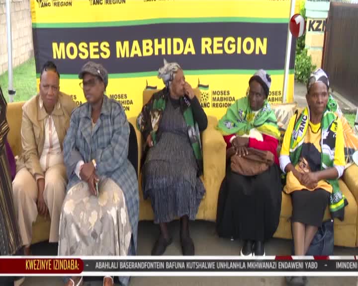 A group of women sit on a yellow couch, their faces turned towards the camera. Behind them, a banner reads "MOSES MABHIDA REGION" in large yellow letters.