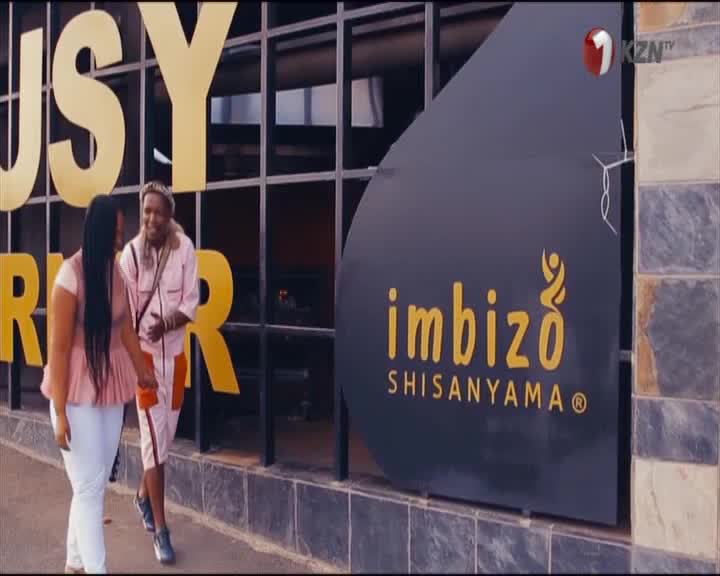 Two women walk past the Imbizo Shisanyama, their conversation animated. The 1KZN TV logo is visible on a sign.