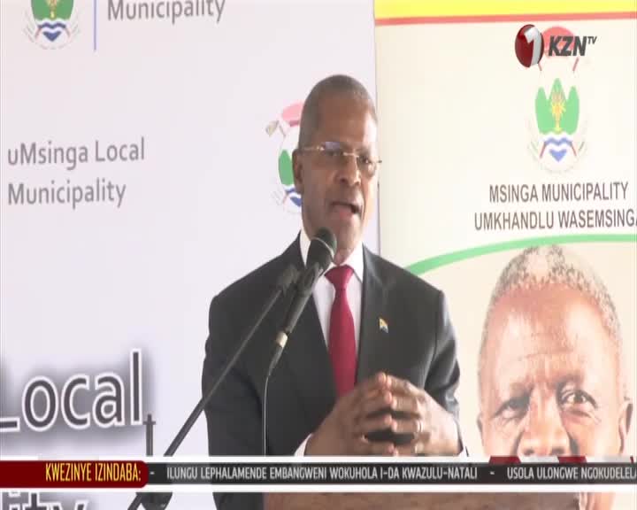A man in a suit and red tie speaks into a microphone at a podium. Behind him, a banner displays the KZN TV logo and the name of the Msinga Municipality. A man in a suit and red tie speaks into a microphone at a podium. Behind him, a banner displays the KZN TV logo and the name of the Msinga Municipality.