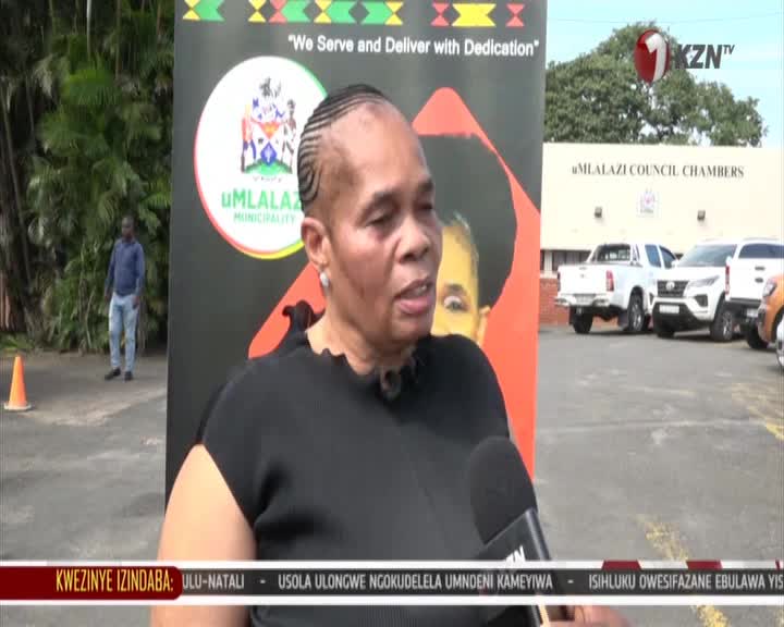 A woman is speaking into a microphone in front of a banner for uMlalazi Municipality. Behind her, vehicles are parked near the uMlalazi Council Chambers.