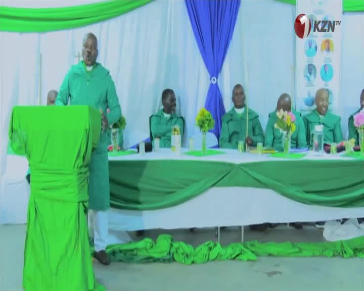 A man in a green robe stands at a podium, speaking to an audience. Seated behind a long table, several other men in matching green robes listen intently.