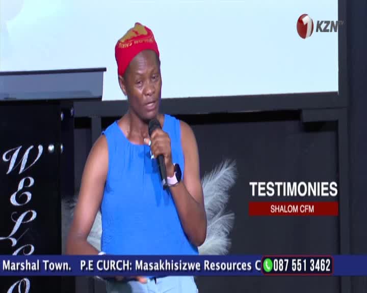 A woman in a blue top and red headwrap holds a microphone, speaking into it. The 1KZN TV logo is visible in the upper right corner.