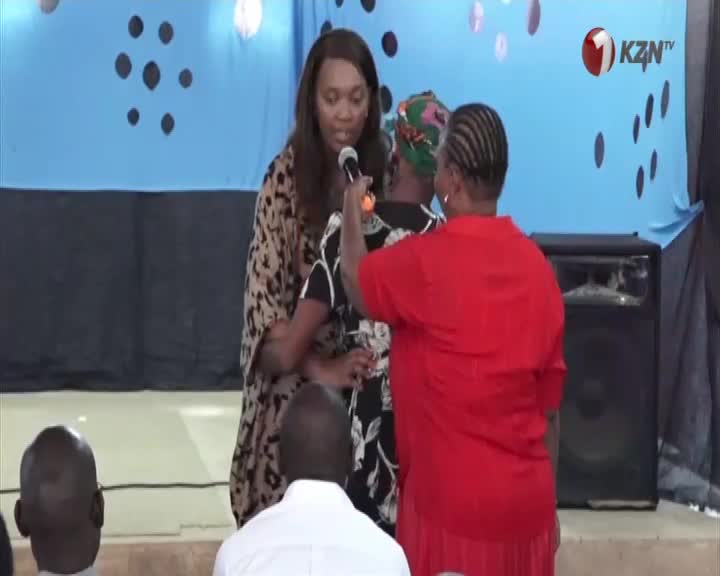 A woman in a leopard-print dress holds a microphone while another woman in a bright red top gently adjusts her headscarf. This moment is being broadcast by KZN TV.