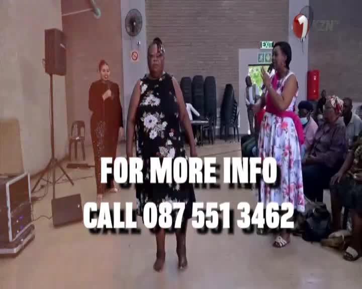 A woman in a floral dress holds a microphone, speaking to a seated audience. Another woman, dressed in black, stands to her left, facing forward.