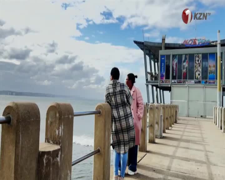 Two women stand on a pier, looking out at the ocean. The KZN TV logo is visible above a building on the pier.