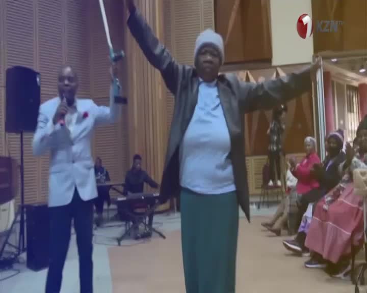 A man in a light suit holds a microphone and a trophy, while a woman in a grey jacket and green skirt raises her arms. The 1KZN TV logo appears in the corner, and an audience watches from the right.