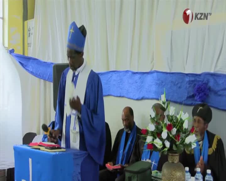A bishop in a blue robe and mitre stands at a lectern, speaking into a microphone. Several other men in religious attire are seated behind him.