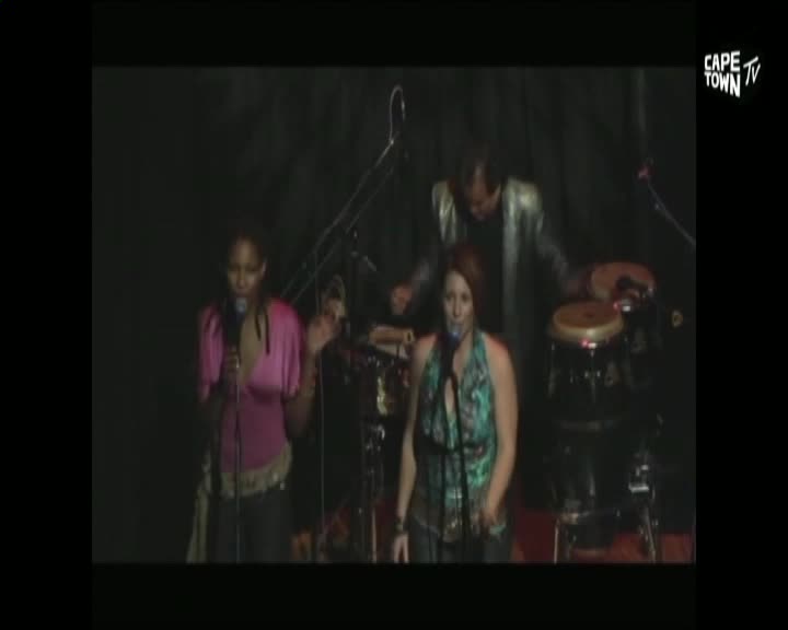 Two women with microphones sing in front of a band, backed by a drummer in a silver jacket. Cape Town TV's logo is in the corner, broadcasting a live performance.
