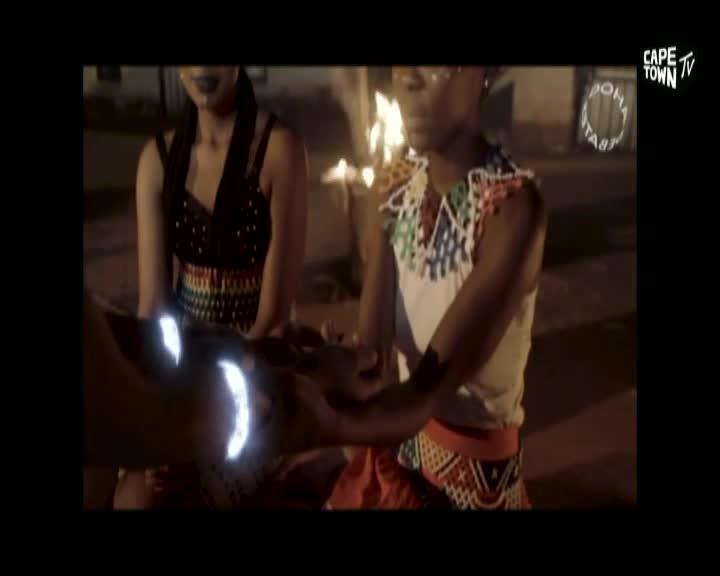 Two women, dressed in colorful beaded attire, are reaching out towards a glowing object. The Cape Town TV logo in the corner suggests this is a performance or demonstration.
