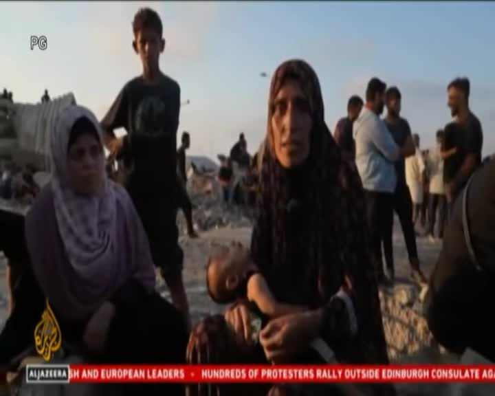 A woman in a dark headscarf holds a child, her face etched with distress. Behind her, others stand and sit amidst rubble, the scene captured on Cape Town TV, Al Jazeera.
