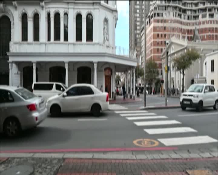 Cars are moving through a Cape Town intersection, with a white sedan turning right. Pedestrians are visible on the sidewalks, and the architecture suggests a historic district.
