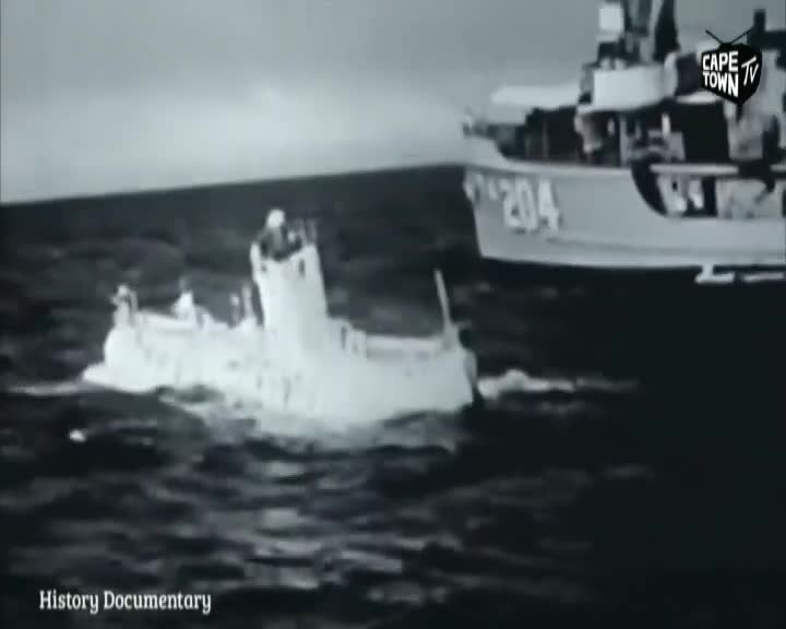A small, white vessel churns through the water, leaving a foamy wake as it moves.  Nearby, a larger ship, marked with the number "204", looms in the background, likely part of the Cape Town TV history documentary.
