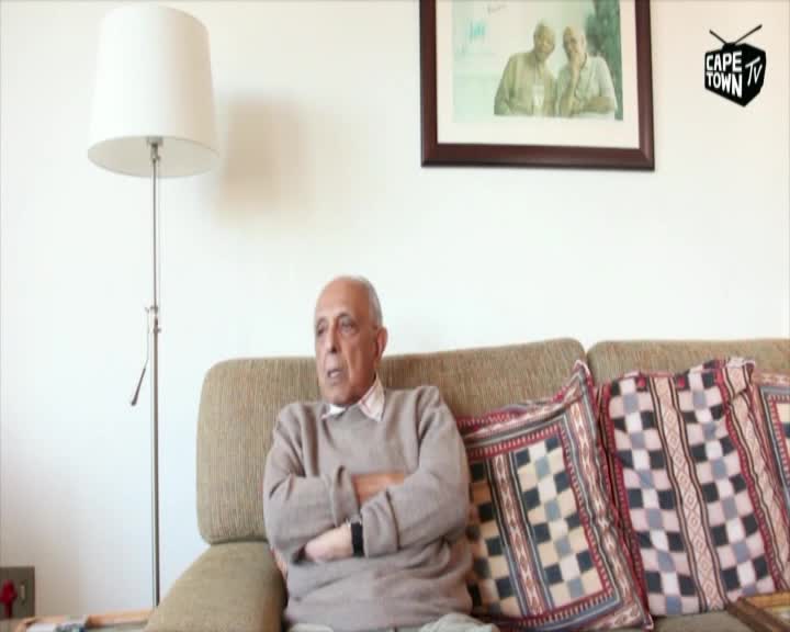 An older man sits on a couch, arms crossed, speaking to someone off-camera. The Cape Town TV logo is visible, and a framed photo hangs above him.
