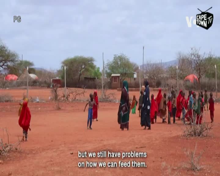 Children in bright red and blue clothing mill about on the dusty ground, while a group of women stand nearby. A Cape Town TV logo is in the corner, and the caption mentions problems with feeding.
