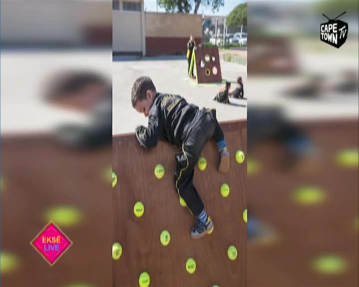 A young boy in a black tracksuit is scaling a wooden wall dotted with tennis balls. He's focused, inching his way up, with Cape Town TV's logo in the corner.
