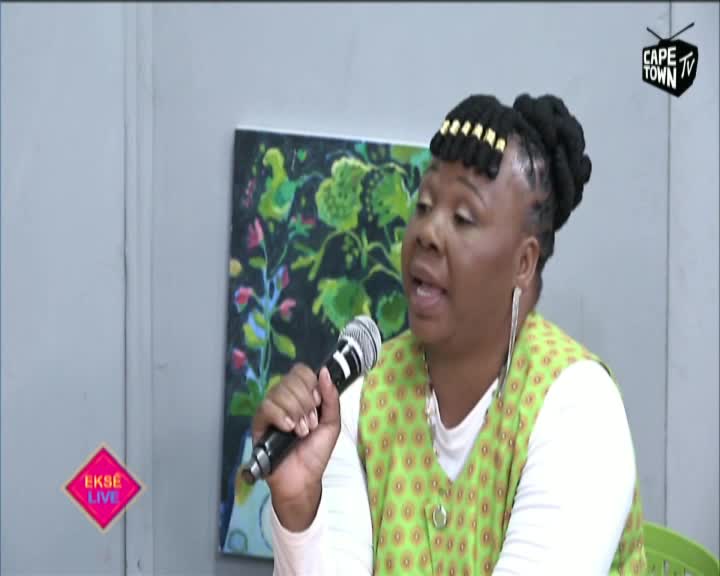A woman in a patterned vest speaks into a microphone, her eyes focused as she addresses the camera on Cape Town TV. Behind her, a colorful painting hangs on the wall, and the "Ekse Live" logo glows in the corner.
