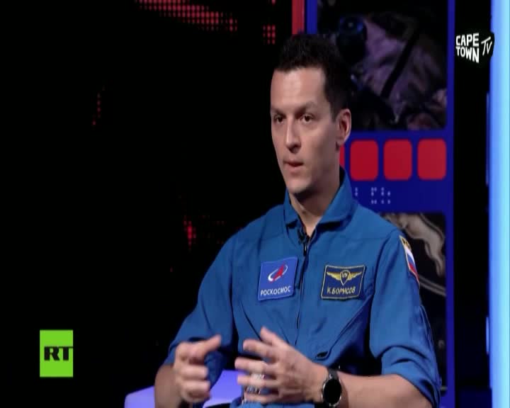 A man in a blue uniform, likely a cosmonaut, gestures with his hands while speaking on Cape Town TV. The RT logo is visible in the corner, and the backdrop is a dark, abstract design.
