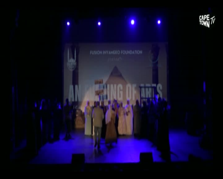 On a stage lit with blue spotlights, a group of people in flowing robes and suits stand before a screen advertising "An Opening of Arts" by the Fusion Inyamazeko Foundation, as broadcast by Cape Town TV. A man in a tan jacket walks towards the center of the stage.
