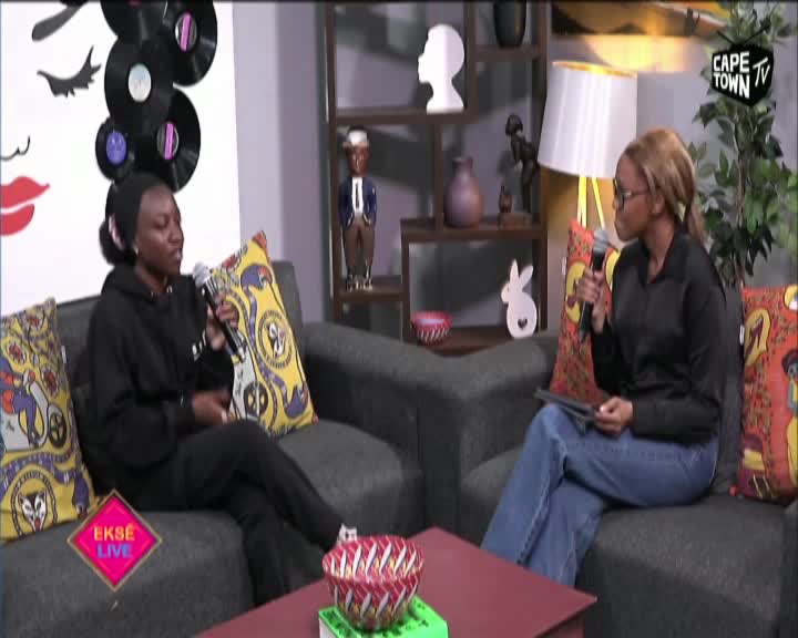 On Cape Town TV, two women sit on a couch, each holding a microphone. The woman on the left, dressed in black, is speaking, while the other, in a black shirt and jeans, listens intently.
