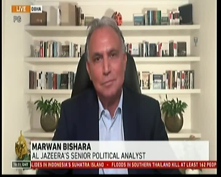 A man in a suit jacket sits in front of a bookshelf, speaking on a screen with the Al Jazeera logo in the bottom left corner. The lower third of the screen displays his name and title, with news headlines scrolling below.
