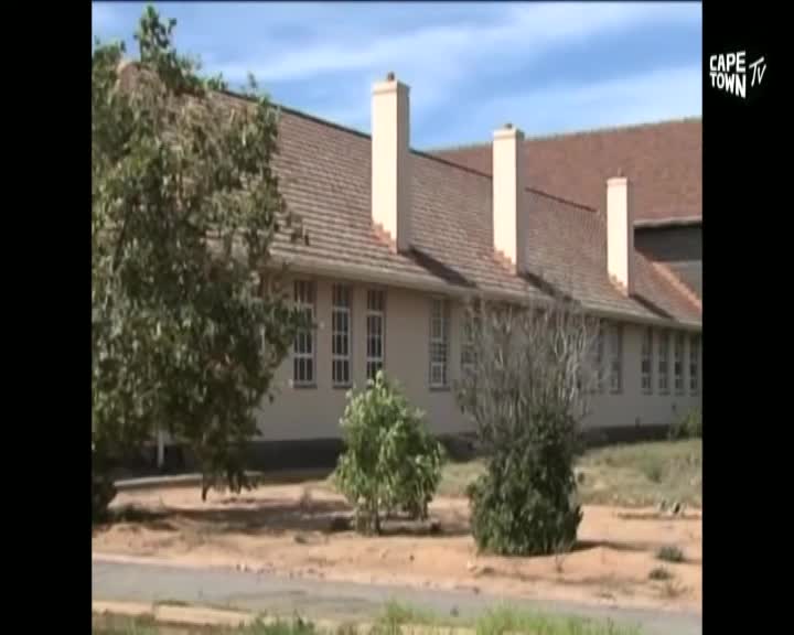 A long, low building with a tiled roof and tall chimneys stretches across the frame. The Cape Town TV logo sits in the corner, and the scene is framed by trees and a bit of dry, sandy ground.
