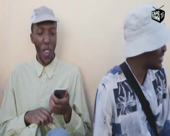 A man in a light green shirt is holding a phone, speaking. Beside him, another man in a patterned shirt is partially visible, and the Cape Town TV logo is on the screen.
