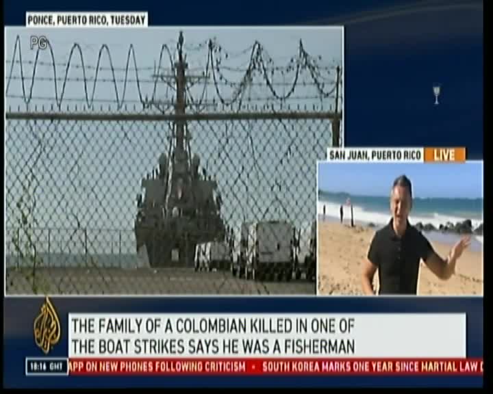 Behind a chain link fence topped with barbed wire, a large ship sits at the dock. A reporter in San Juan, Puerto Rico, gestures towards the beach while discussing the story.
