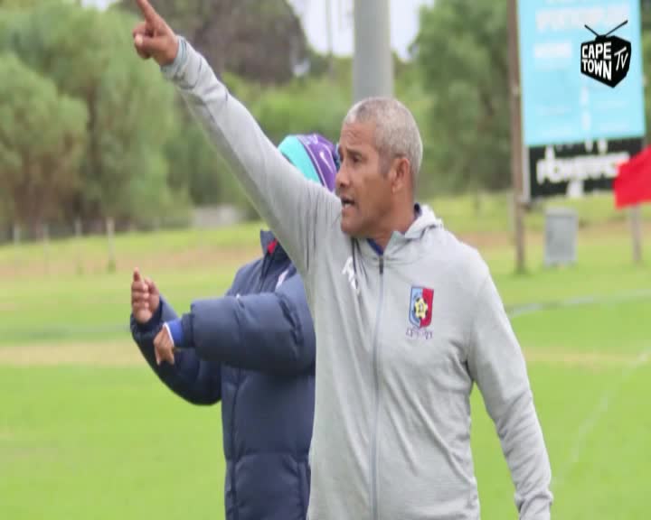 The coach, wearing a grey jacket, is pointing and shouting, his arm extended towards the field. A Cape Town TV logo is visible as he directs the team.
