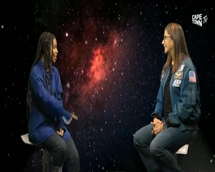 Two women sit facing each other on stools, engaged in conversation against a backdrop of stars, as seen on Cape Town TV. The woman on the left gestures with her hands while the other listens attentively, wearing a jacket with an American flag patch.
