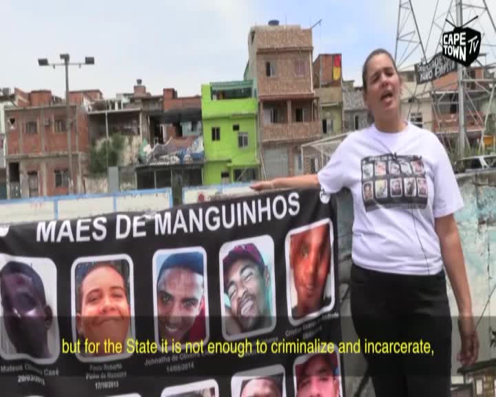 A woman stands next to a banner displaying photos of young men, her voice rising as she speaks. Behind her, a dense neighborhood of colorful buildings climbs the hillside.